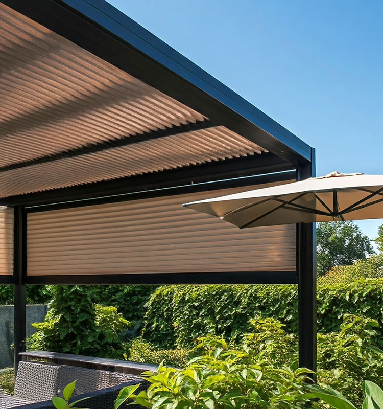 Modern outdoor patio with black pergola frame and retractable beige awning, surrounded by lush garden landscaping and blue sky