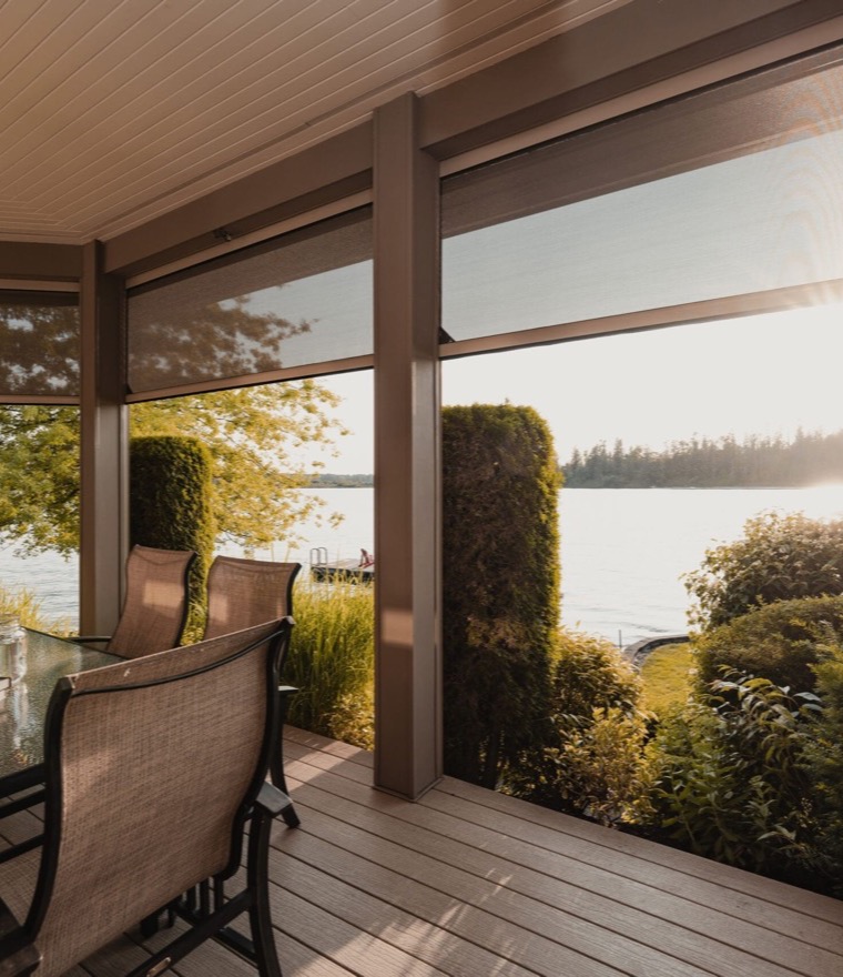 Screened porch with motorized retractable screens lowered around a glass dining table, overlooking a lake at sunset