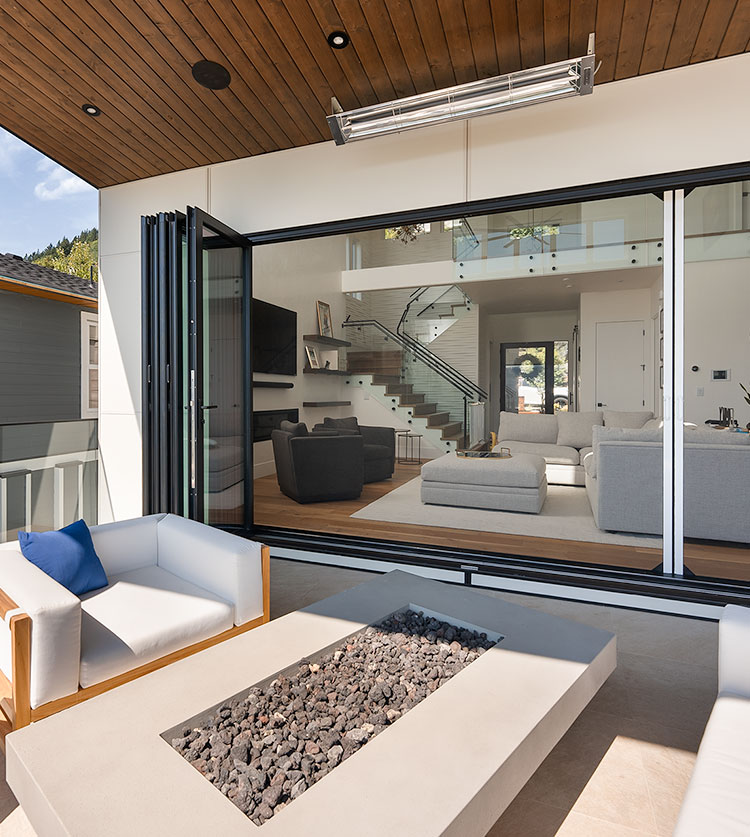 Modern covered patio with folding glass doors, white outdoor furniture, and built-in fire pit looking in on a contemporary living room