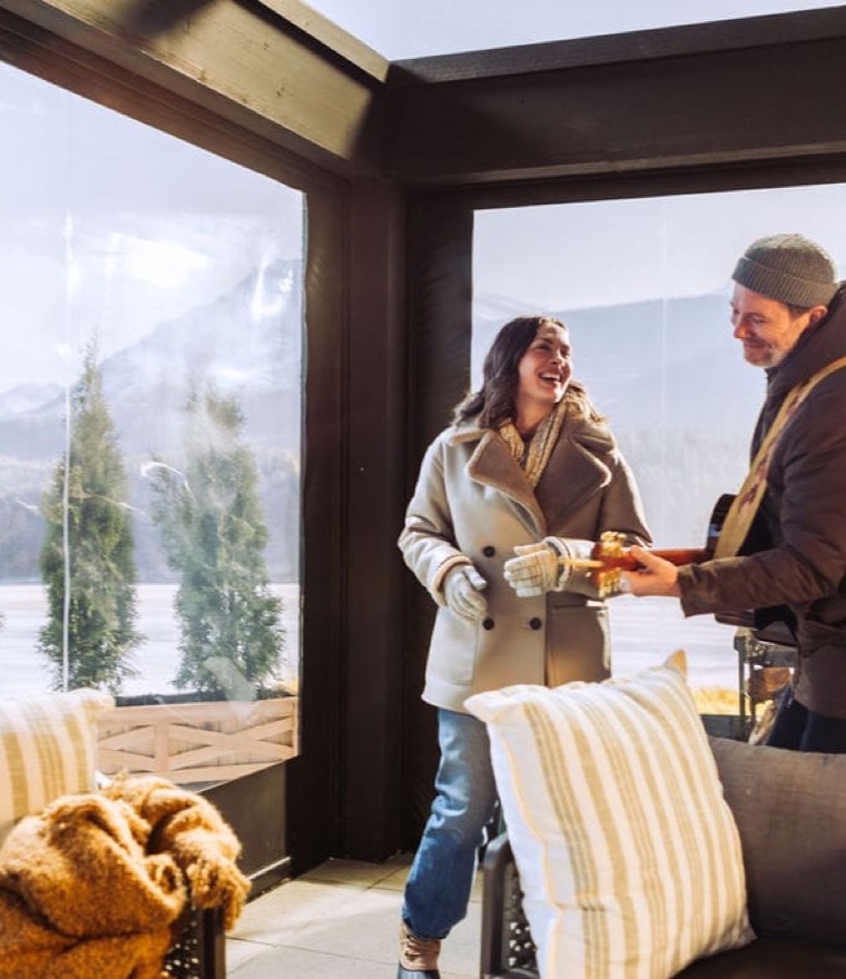 Couple in winter coats sharing a warm drink on a luxury-vinyl-enclosed patio overlooking a frozen lake and mountains