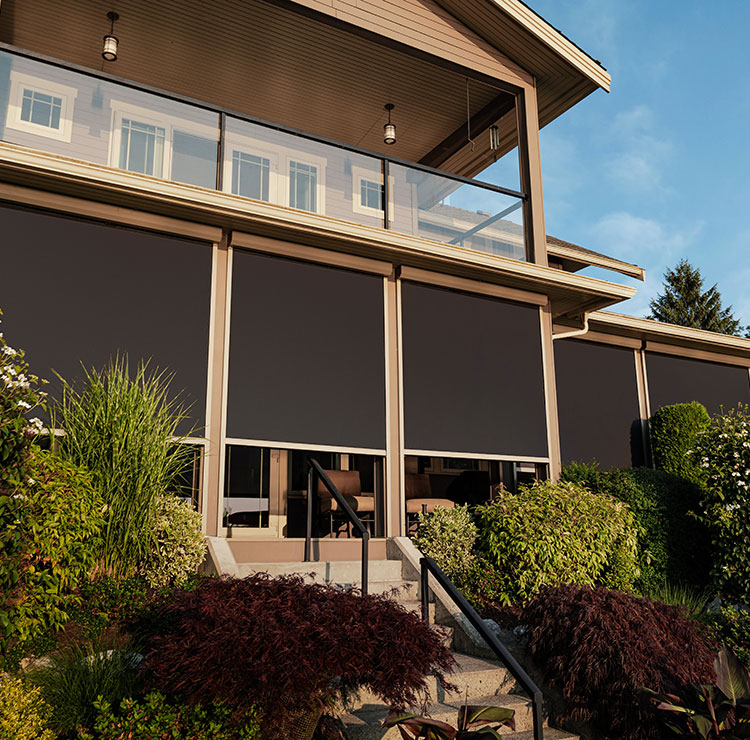 Modern home exterior with dark motorized screens lowered on covered patio, featuring landscaped garden and glass railings above