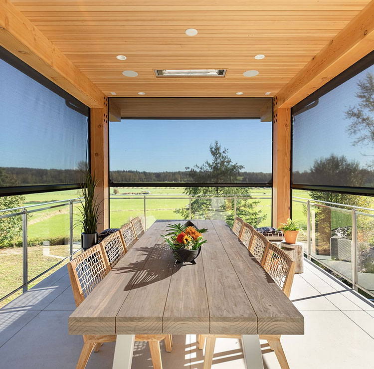 Covered dining area with wooden table and motorized screens, offering panoramic views of green landscape