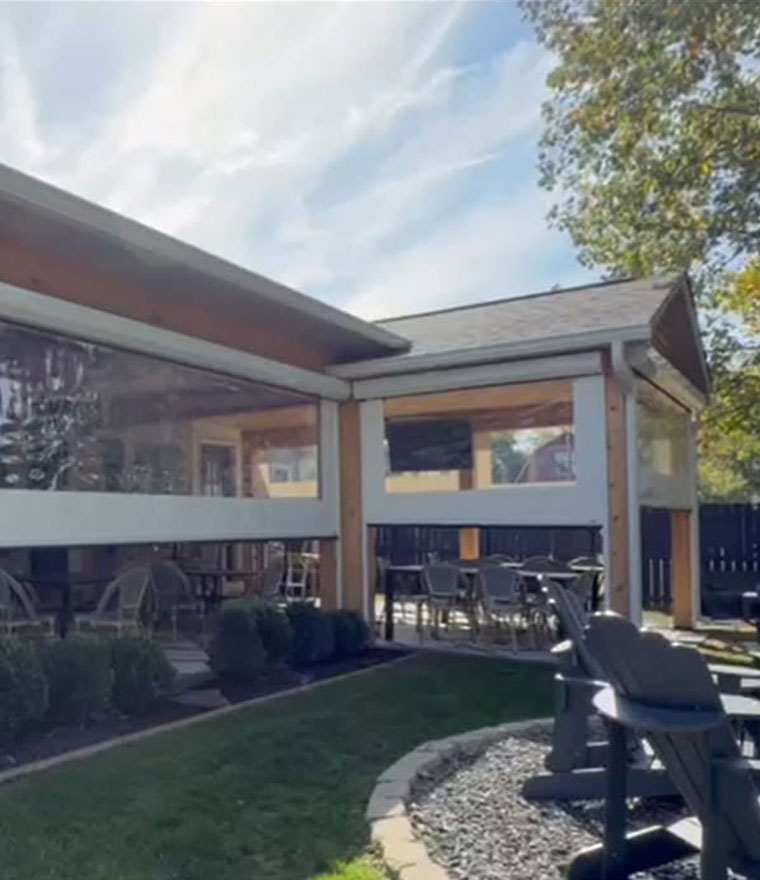 Exterior of a commercial building with large clear vinyl retractable screens enclosing a covered patio, dining chairs and tables visible through the transparent panels