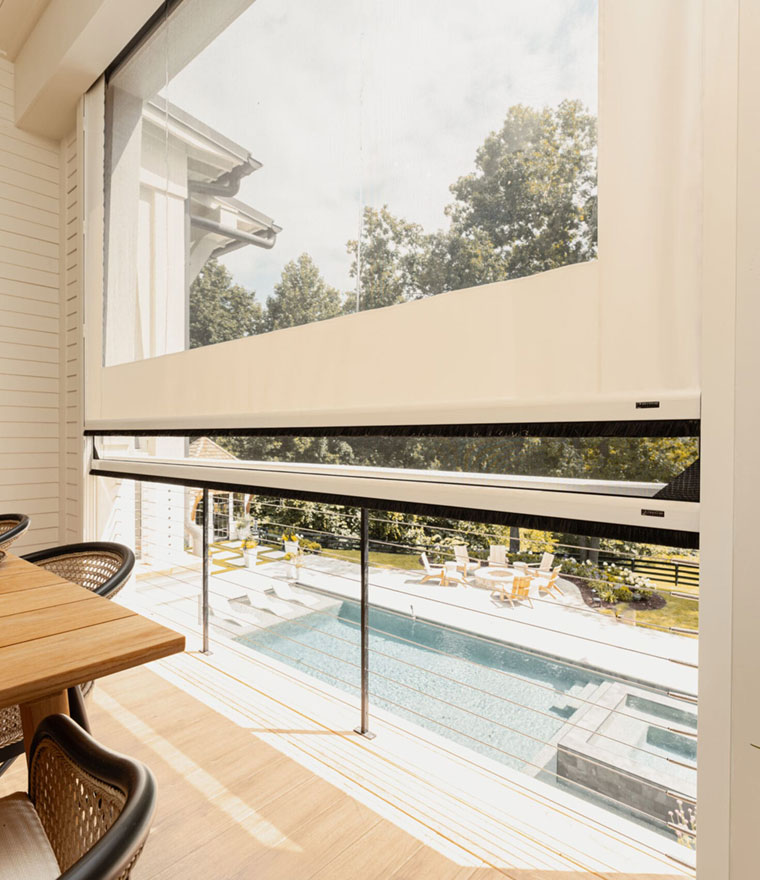 Interior view of a screened balcony with two retractable screens partially deployed over a glass-railed deck, pool terrace and outdoor furniture visible below