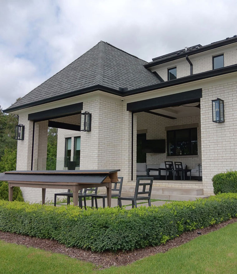 Exterior of a white brick home with black retractable screens installed under a hip-roof covered patio, outdoor dining table and bench in the foreground
