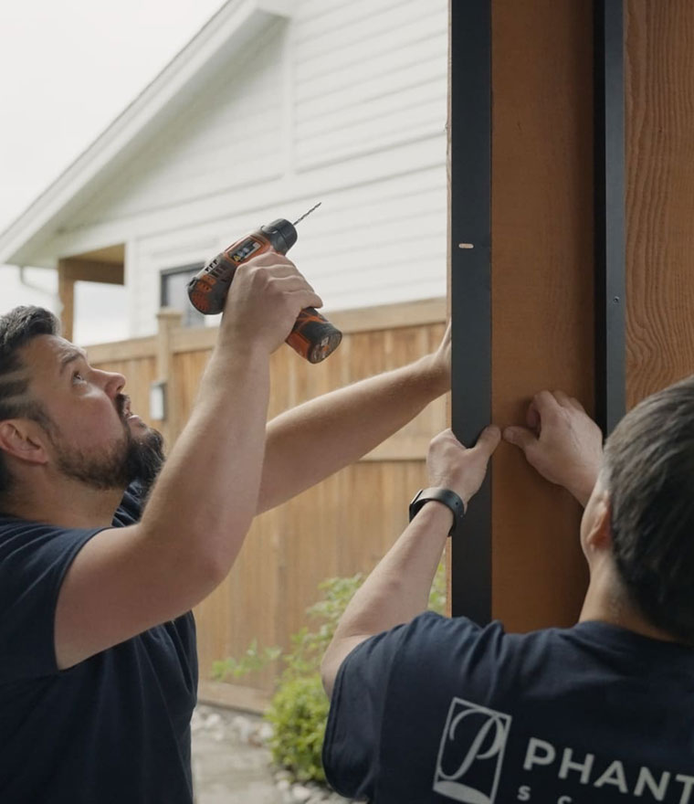 Two Phantom Screens installers mounting a screen side track on a wood-framed opening, one using a power drill and the other steadying the track