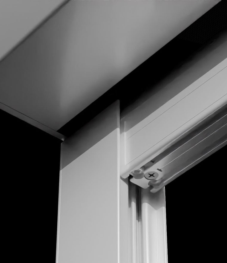 Black and white close-up of the screen housing mounted at a wall and ceiling corner, showing the aluminium track profile and a visible Phillips head adjustment screw