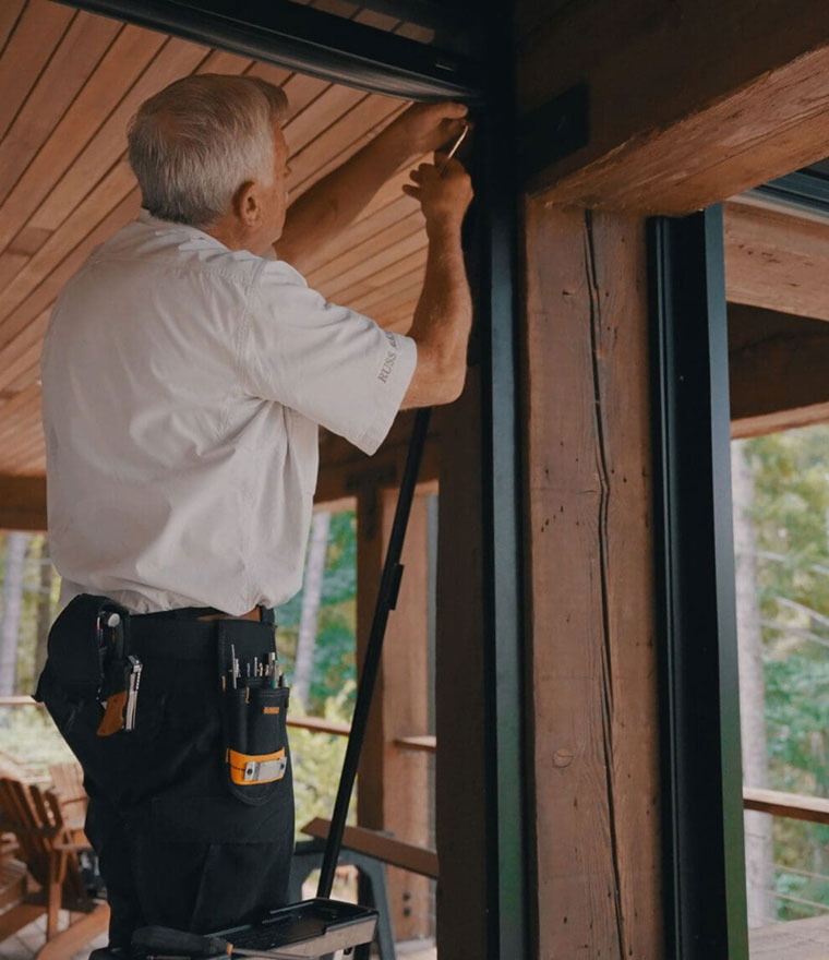 Installer on a ladder mounting a motorized screen housing on a rustic wooden covered porch
