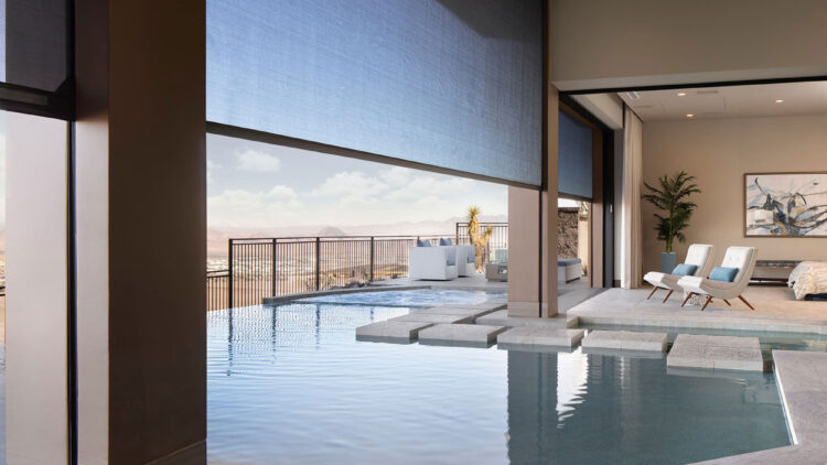 Luxury indoor-outdoor pool area with blue retractable shades, stone flooring, and modern lounge chairs overlooking a desert landscape